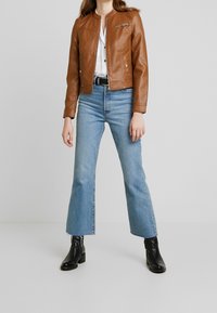 Brun læderbikerjakke med tætsiddende design, lynlåslomme og knaplukning med trykknapper. Kombineret med vide blå jeans og sorte støvler.