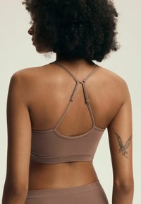 Brązowy ribbed braletka z regulowanymi cienkimi ramiączkami. Posiada krzyżowy design na plecach oraz bezszwowe krawędzie dla większego komfortu. Zawiera widoczne tatuaż na ramieniu.