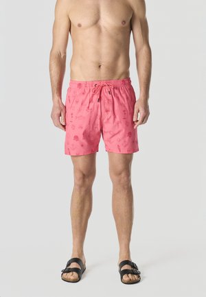 Hombre sin camisa con pantalones cortos de baño rosas con estampados náuticos y sandalias negras con hebilla, de pie contra un fondo claro y liso.