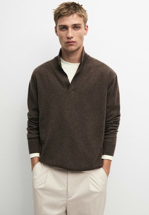 Giovane uomo che indossa un maglione marrone sopra una camicia bianca con pantaloni beige chiaro, in piedi con le mani in tasca davanti a uno sfondo semplice.