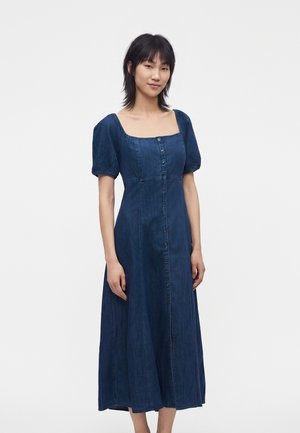 Vrouw staand in een blauwe denim midi-jurk met korte pofmouwen, vierkante halslijn en knopen aan de voorkant, tegen een effen lichte achtergrond.