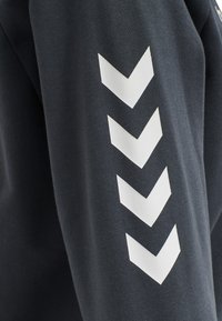 Czarna bluza z białymi wzorami chevron wzdłuż rękawów. Wykonana z miękkiego materiału o gładkiej fakturze. Prosty i nowoczesny design.