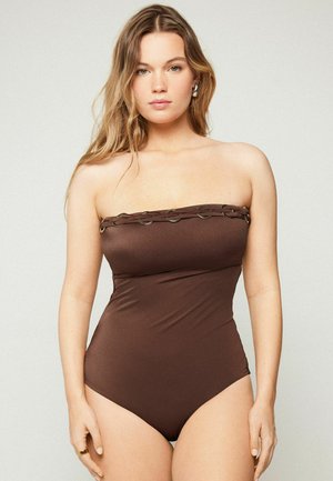 Femme portant un maillot de bain une pièce marron sans bretelles avec une bordure tressée et des anneaux métalliques, debout devant un fond clair uni.