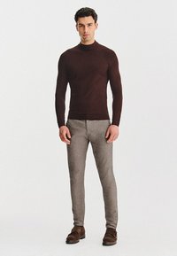 Brązowy dopasowany sweter z wysokim kołnierzem, sparowany ze światłymi szarymi spodniami o slim fit i brązowymi butami. Gładki materiał z minimalną fakturą.