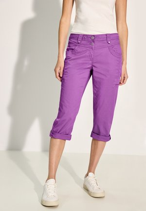 Violette kurze Hosen mit zwei vorderen Taschen, umgeschlagenen Säumen und glatter Textur. Kombiniert mit weißen Sneakers.