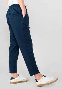 Pantalones joggers de color azul marino con cintura elástica, bolsillos laterales y un diseño ajustado, combinados con zapatillas blancas con suelas marrones.