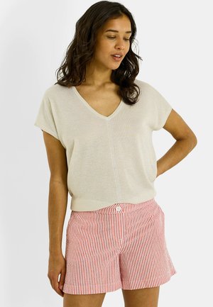 Mujer con tono de piel medio y cabello oscuro, vistiendo una camiseta de cuello en V de color beige claro y pantalones cortos a rayas rojas y blancas, de pie con una mano en la cadera.