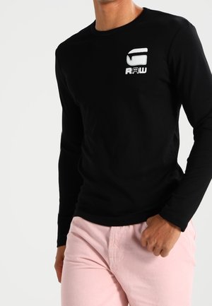 Man wearing black long-sleeve shirt with white logo on chest and light pink pants against plain white background.