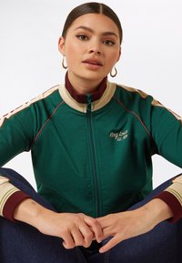 Jonge vrouw met bruin haar in een groene ritsjack met borduursel "King Louie Est. 1984", zittend met gekruiste benen, handen samengevouwen, met gouden ringoorbellen.