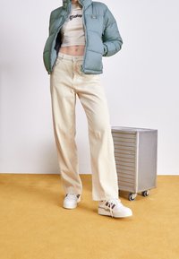 Puffed mint-green jacket, white cropped shirt, cream corduroy wide-leg pants, and chunky white sneakers on a yellow background. Silver suitcase nearby.