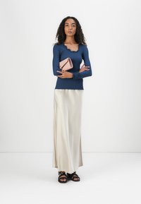 Blusa de manga longa azul-marinho com textura em ribetes e decote escallopado, combinada com uma saia maxi em cetim creme e sandálias pretas. A segurá uma clutch rosa-claro.