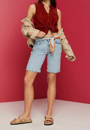 Jeansshorts med ljus tvätt, matchat med en röd ärmlös topp och en beige jacka, med en flerfärgad scarf knuten i midjan.
