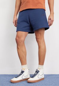 Shorts bleu marine en tissu doux, avec une taille élastique et un discret écusson logo. Portés avec des baskets montantes blanches et des chaussettes mi-mollet.