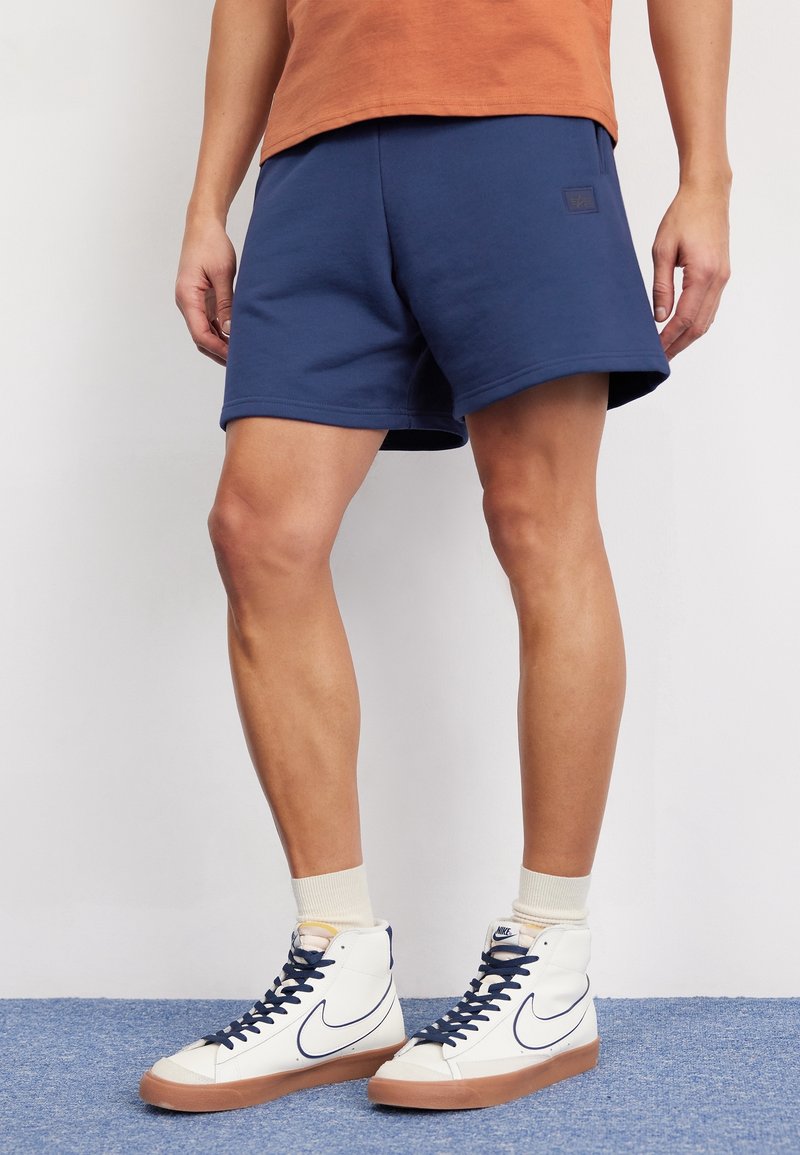 Shorts bleu marine en tissu doux, avec une taille élastique et un discret écusson logo. Portés avec des baskets montantes blanches et des chaussettes mi-mollet.