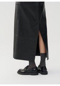 Jupe en cuir noir avec une fente sur le côté, associée à des chaussures noires brillantes à enfiler et des chaussettes opaques gris foncé. Texture lisse, design moderne.