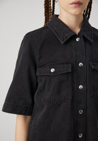 Svart denimskjorta med korta ärmar, framträdande krage, två bröstfickor och silverfärgade tryckknappar, med en texturerad och något urblekt yta.