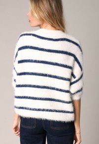 Un pull blanc en laine moelleuse avec des rayures horizontales bleu marine, des épaules tombantes et une coupe large. Le matériau semble doux et texturé.