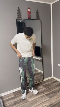 Bijela majica s kratkim rukavima s zelenim logotipom, uparena s uzorkanim hlačama u tamnozelenoj, ljubičastoj i bijelim dijamantima, nošena s bijelim tenisicama.