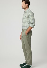 Camisa a cuadros verde claro con botones y mangas enrolladas; pantalones ajustados beige; zapatillas verdes con detalles en blanco. El modelo está de pie de lado.