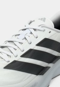 Άσπρο αθλητικό παπούτσι με υφή στην επάνω επιφάνεια, διαθέτει τρεις μαύρες ρίγες, άσπρα κορδόνια και στρογγυλεμένο σχεδιασμό στη φτέρνα.