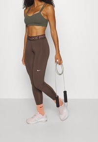 Kobieta w brązowych legginsach Nike, oliwkowym biustonoszu sportowym, różowych skarpetkach i trampkach trzymająca skakankę z czarnymi uchwytami na jednolitym tle.