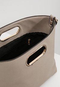 Beige laukku, jossa on tekstuuri, kultasävyiset yksityiskohdat ja kaksinkertaiset leikkuukahvat. Sisätilassa on musta vuori ja vetoketjullinen tasku.