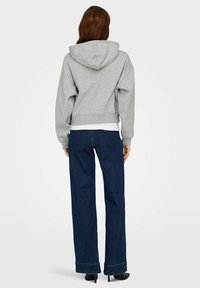 Femme aux longs cheveux bruns portant un sweat à capuche gris, un jean large bleu foncé et des escarpins noirs, tournant le dos sur un fond clair uni.