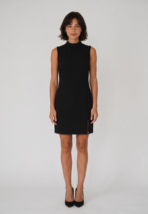 Donna con capelli castani corti che indossa un abito nero senza maniche con uno spacco laterale e tacchi alti neri, in piedi davanti a uno sfondo chiaro e uniforme.