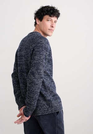 Tamno plavi pleteni pulover s teksturiranim uzorkom, dugih rukava i okruglim izrezom; vidljiva meka tkanina. Bijeli detalji kroz cijeli pulover.