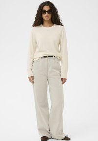 Sieviete ar cirtainiem matiem, valkājot melnus saulesbrilles, krēmkrāsas džemperi, bēša platās bikses, melnu jostu un brūnas kurpes, nostājusies pret baltu fona.