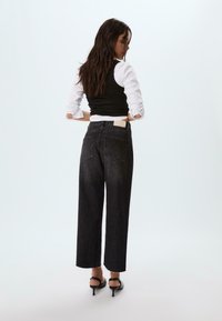 Jeans neri a vita alta e gamba ampia con orlo grezzo, tasche posteriori e un'etichetta di colore chiaro sulla cintura. Il modello indossa un top aderente nero.