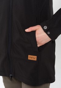 Chaqueta exterior negra con cremallera de longitud completa, bolsillos laterales y una etiqueta de cuero. Textura suave con detalles de botones en los puños.
