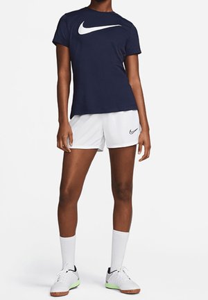 Osoba ubrana w granatowy t-shirt Nike, białe szorty Nike, białe skarpety za kostkę i białe sneakersy, stojąca na tle jednolitego tła.