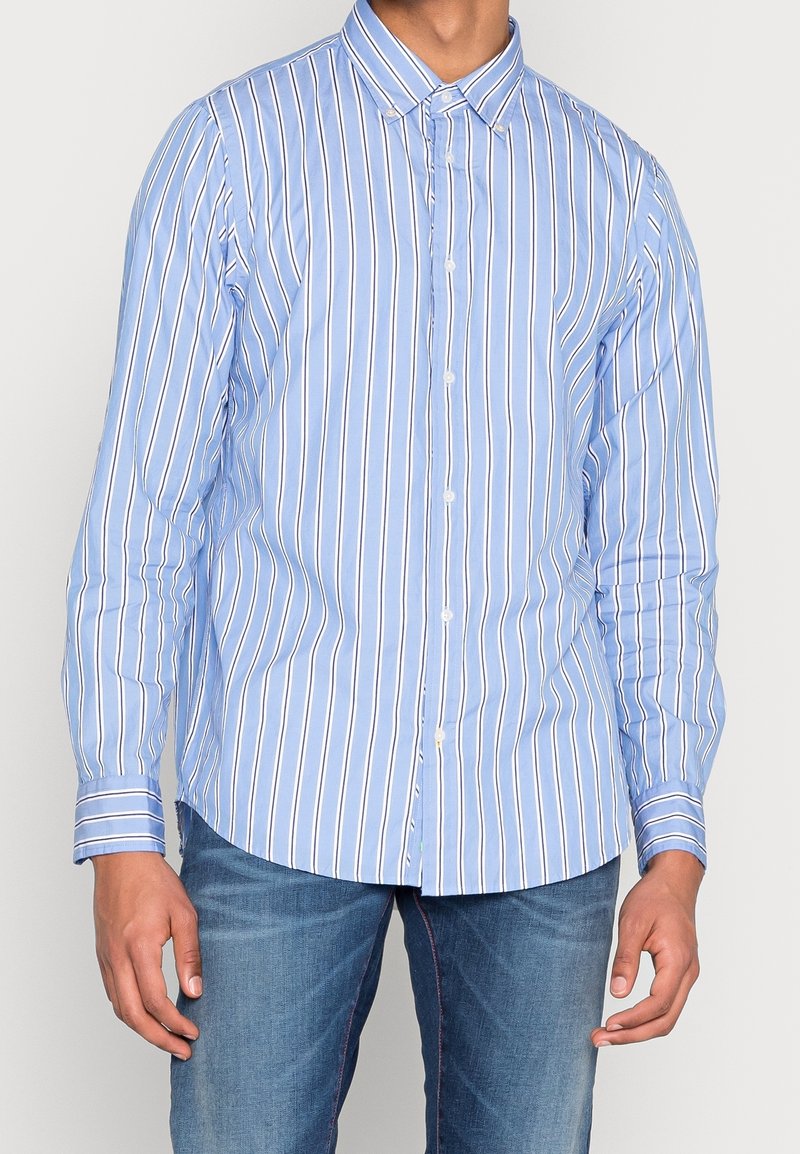 Lyseblå skjorte med knapper, der har lodrette hvide og navyblå striber. Den har lange ærmer, en spids krave og en buet kant.