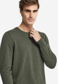 Grön långärmad tröja med rund halsringning och ribbade muddarna, gjord av ett mjukt, texturerat tyg. En man justerar sin krage.