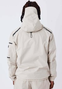 Giacca isolante beige chiaro con cappuccio, caratterizzata da una striscia nera di accento sulle spalle e sui polsini, e polsini elasticizzati per una vestibilità aderente.