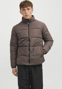 Giubbotto imbottito marrone con colletto alto, zip frontale e due tasche laterali; presenta cuciture orizzontali e polsini a costine. Pantaloni neri sotto.