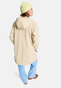 Beige Kapuzenjacke mit lässigem Schnitt, kombiniert mit hellblauen Jeans. Verfügt über eine Tasche mit pinkem Akzent und einer strukturierten gelben Mütze.