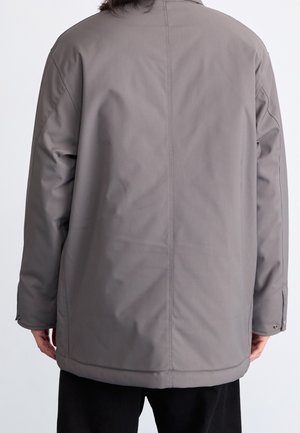 Person iført en langærmet, grå jakke set bagfra, med sorte bukser og hænder afslappet ved siderne.