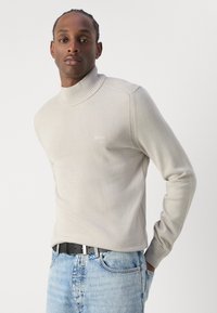 Suéter de cuello alto beige en tejido de punto con cuello y puños acanalados. Presenta un sutil logotipo en el pecho. Combinado con pantalones vaqueros azul claro y cinturón negro.