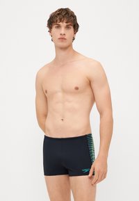 Fekete úszónadrág, testhez simuló kialakítással, az oldalán textúrázott zöld mintával és egy kicsi Speedo logóval az alján.