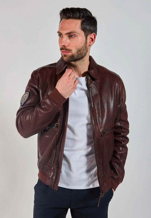 Veste en cuir marron avec fermeture éclair, poignets côtelés et un écusson circulaire sur la manche gauche. Portée par-dessus un t-shirt blanc.