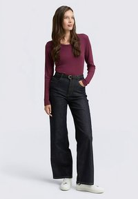 Langärmeliges burgunderfarbenes Oberteil, kombiniert mit hoch taillierten, weit geschnittenen dunklen Jeans und weißen Sneakers. Die Jeans haben einen Gürtel und Taschen.