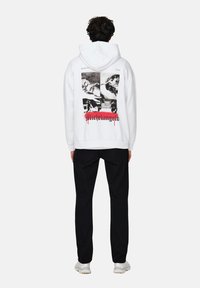 Sudadera blanca con estampados gráficos negros en la parte posterior, que presenta detalles artísticos y la palabra "Michelangelo" en rojo. Lleva puestos pantalones negros.