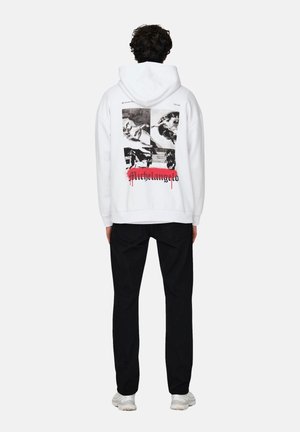 Sudadera blanca con estampados gráficos negros en la parte posterior, que presenta detalles artísticos y la palabra "Michelangelo" en rojo. Lleva puestos pantalones negros.