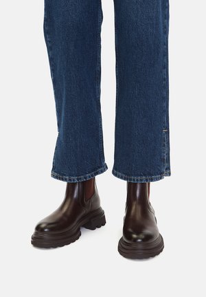 Brązowe skórzane botki za kostkę z elastycznymi panelami po bokach i grubą, teksturowaną podeszwą, noszone z niebieskimi dżinsami o skróconym kroju.