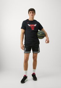 Black T-shirt with red bull graphic, black shorts with white accents, black sneakers with pink details, and holding a black basketball.