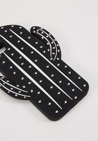 Coque de téléphone en forme de cactus noir avec des motifs et des lignes blanches en flèche. Fini texturé, découpes pour l'appareil photo et les boutons. Matériau thermoplastique.