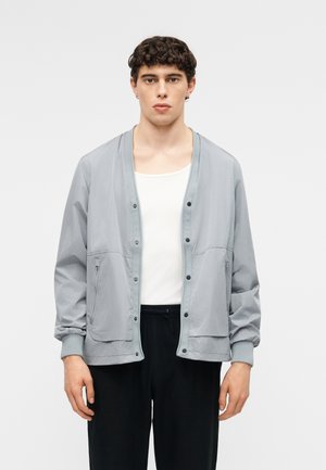 Ung mand med kort krøllet hår iført en lysegrå knappet jakke, hvid tanktop og sorte bukser foran en ensfarvet baggrund.