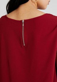 Czerwony strój o teksturowanej powierzchni, z zaokrąglonym dekoltem i widocznym metalicznym zamkiem błyskawicznym z tyłu.